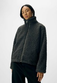 Veste en polaire grise zippée avec un col haut, un extérieur texturé et une coupe droite. Elle possède des manches longues et une fermeture éclair en métal.