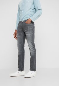 Vaqueros gris ajustados con textura deslavada, cinco bolsillos y cierre de botón metálico, combinados con una camisa de mezclilla azul claro y zapatillas blancas sin cordones.