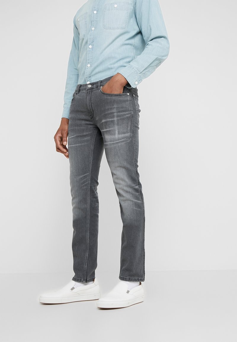 Vaqueros gris ajustados con textura deslavada, cinco bolsillos y cierre de botón metálico, combinados con una camisa de mezclilla azul claro y zapatillas blancas sin cordones.