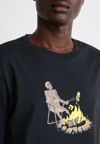 Camiseta de algodón negra con un estampado gráfico de un esqueleto sentado en una silla, asando un perrito caliente sobre un fuego, con llamas amarillas vibrantes.