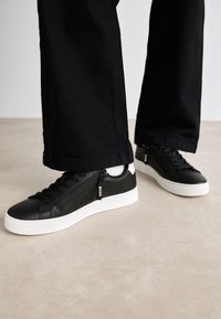 Black leather sneakers with white soles and side zippers, worn with black pants and white socks on a beige floor.