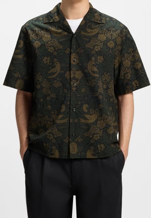 Chemise à manches courtes vert foncé à boutons avec un motif d'oiseaux bruns et de fleurs, portée par-dessus un sous-vêtement blanc.
