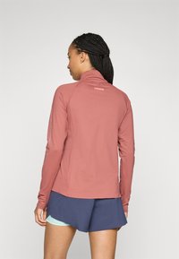 Langärmliges Oberteil mit hohem Kragen aus rosafarbenem Stoff mit glatter Textur. Mit Logo auf der Rückseite und reflektierenden Akzenten an den Ärmeln. Kombiniert mit Shorts.