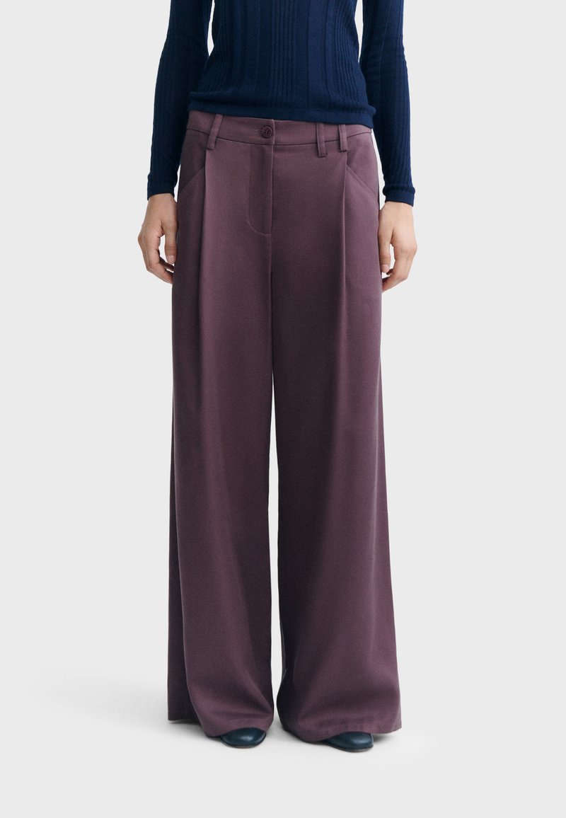 Personne portant un pantalon violet à jambes larges et une chemise à manches longues bleu foncé côtelée, debout devant un fond uni.