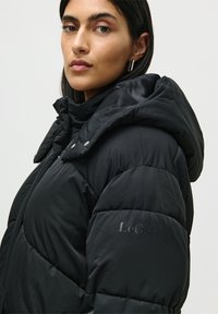 Mujer con cabello oscuro que lleva pendientes de aro de plata y una chaqueta acolchada negra con capucha, mirando ligeramente por encima de su hombro.