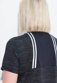 Osoba s rovnými blond bob vlasy, oblečená v černém sportovním tričku s jemnými zlatými linkami a dvěma vertikálními bílými pruhy na zádech.