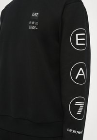 Black sweatshirt with a crew neckline. Features white printed logos: "EA7" and "Emporio Armani." Smooth fabric with ribbed cuffs and hem.