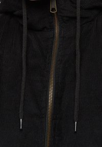 Black corduroy jacket front with brass zipper and black drawstrings featuring metal tips.