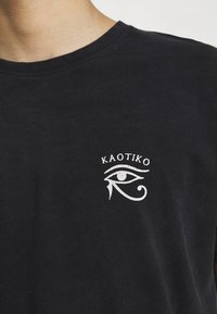 Černé bavlněné tričko s bílou grafikou oka a textem "KAOTIKO" nad ním. Kulatý výstřih, krátké rukávy.