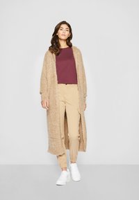 Beige struktureret lang cardigan over en maroon top, parret med beige bukser og hvide sneakers. Blødt, fyldigt materiale med løs pasform.