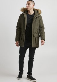 Maslinasto zelena parka sa kapuljačom od krzna, patent zatvaračem sprijeda, više džepova i opuštenog kroja, uparen s crnim hlačama i tenisicama.