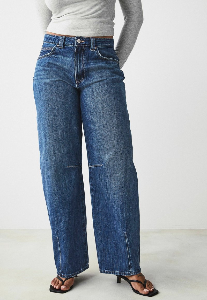 Jeans in denim blu a gamba larga con vita alta, dotati di leggera usura e sottili dettagli di cucito. Abbinati a sandali neri con tacco.