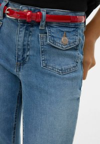 Modré džínové kalhoty s kapsou na klopu na knoflíky a lesklým červeným páskem, nošené s černým topem.