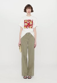 T-shirt bianca con una grafica floreale rossa, abbinata a pantaloni larghi verde oliva. La modella tiene in mano una pochette dorata e indossa sandali neri.