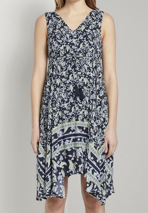 Robe sans manches bleu marine à motif floral vert et blanc, col en V, détail noué à la taille, et ourlet asymétrique portée par une personne debout.