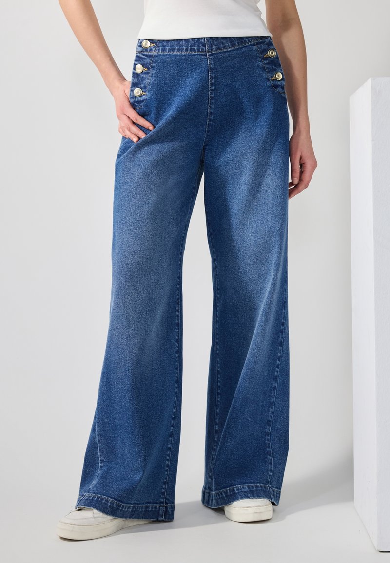 Jean en denim bleu à jambes larges avec des boutons dorés à la taille, porté avec des baskets blanches et un haut blanc, présenté de la taille aux pieds.