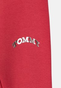 Crveni pamučni materijal s zakrivljenim "TOMMY" logom u metalik srebrnoj boji. Glatka tekstura bez uzoraka ili dodatnih detalja.