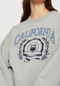 Sudadera gris de manga larga, con gráficos impresos en azul y marino. El diseño incluye "California" y un emblema con ramas de laurel.