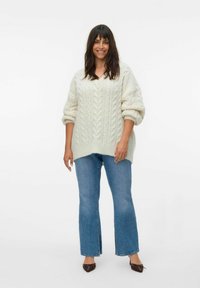 Suéter de punto grueso en color crema y de gran tamaño, con un escote en V profundo, combinado con jeans acampanados de color azul claro y zapatos marrones de punta.