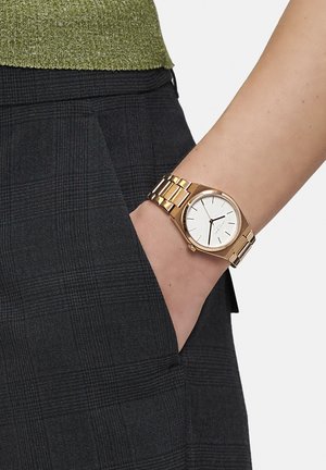 Guldtonet ur med en rund hvid urskive og minimalistiske sorte timeindikatorer, parret med en linksarmbånd, båret på en hånd i en lomme.