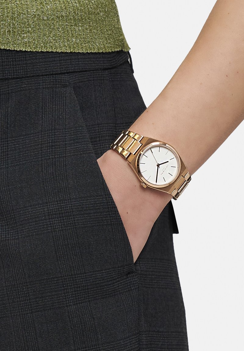 Guldfärgad klocka med en rund vit urtavla och minimalistiska svarta timmarkörer, tillsammans med ett länkarmband, bärs på en hand i fickan.