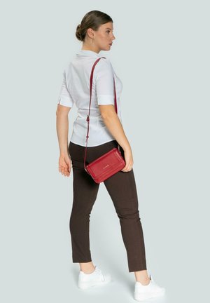 Bolso cruzado rojo con acabado texturizado, que cuenta con una correa larga. El modelo lleva una camisa blanca y pantalones marrones, con zapatillas blancas. Diseño simple.