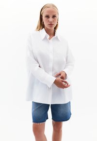 Camicia bianca con colletto classico e maniche lunghe, abbinata a pantaloni shorts in denim blu con bordi sfrangiati.