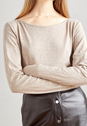 Vrouw draagt een beige top met lange mouwen en een boothals, die haar armen kruist, gecombineerd met een hooggesloten zwarte lederen rok met zilveren knopen.
