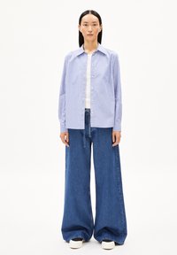 Camicia button-up a righe in azzurro chiaro e bianco, abbinata a jeans a gamba larga blu scuro. Il modello indossa sneakers bianche e si trova su uno sfondo semplice.