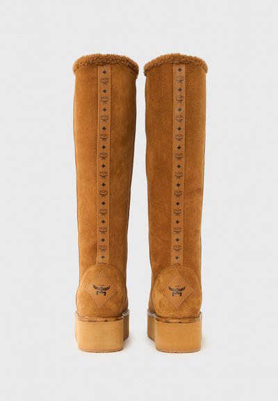 Bottes hautes en daim marron avec doublure en fourrure, comprenant une sangle décorative avec des logos et une semelle plateforme épaisse. Fini texturé sur la tige.