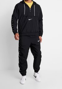 Man i svart Nike-träningsdräkt med vita logotyper och vita Nike-sneakers, som står mot en enfärgad ljus bakgrund.