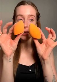 Femme aux longs cheveux tenant deux éponges à maquillage orange près de son visage, montrant leur forme et leur taille sur un fond uni.