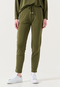 Olijfgroene joggingsbroek, elastische tailleband met trekkoord, slim fit, zachte stof, zijkanten met strepen, gecombineerd met witte sneakers.