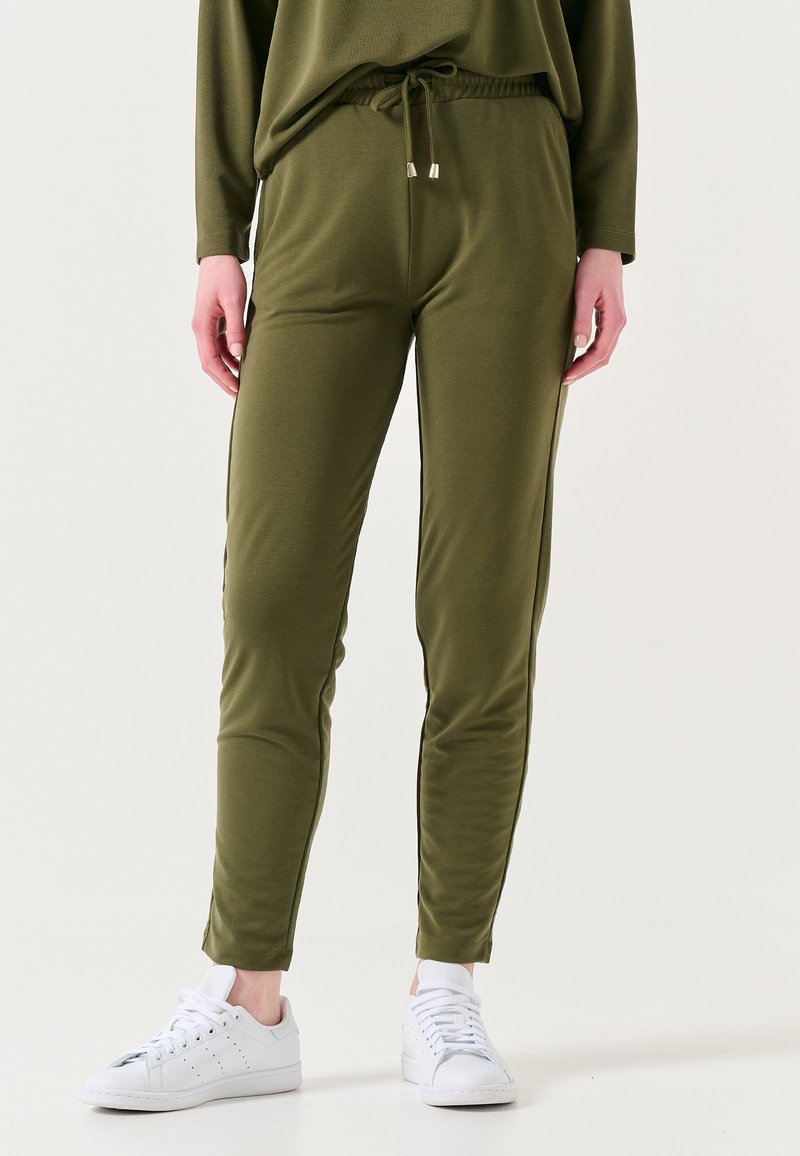 Olijfgroene joggingsbroek, elastische tailleband met trekkoord, slim fit, zachte stof, zijkanten met strepen, gecombineerd met witte sneakers.
