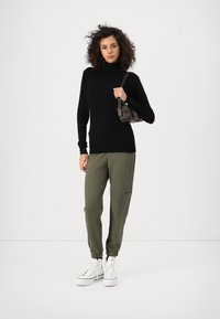 Jersey negro de cuello alto, pantalones jogger oliva con bolsillos y zapatillas blancas. La modelo sostiene un pequeño bolso marrón. Fondo blanco liso.