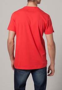 Camiseta de algodón roja con mangas cortas, escote redondo y dobladillo recto, llevada con pantalones de mezclilla azules. Sin marcas ni patrones visibles.