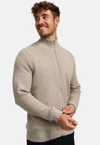 Beige gebreide ritsvest met een gestructureerd patroon, hoge kraag en geribbelde manchetten. Kenmerkt zich door een contrasterende rits en een getailleerde pasvorm.
