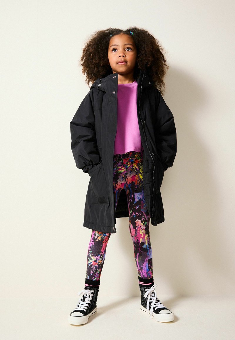 Sort isoleret frakke, lyserød cropped sweatshirt, multikolorede leggings med abstrakte mønstre og sorte sneakers.