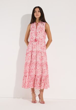 Frau trägt ein ärmelloses, pinkfarbenes Maxikleid mit Blumenmuster und vorderer Schleife sowie pinkfarbene Sandaletten mit hohen Absätzen vor einer schlichten weißen Wand.