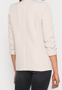 Jasno beżowa marynarka z podwiniętymi rękawami, charakteryzująca się dopasowanym krojem i gładką strukturą. Zawiera dwie kieszenie boczne i pojedynczy szew z tyłu.