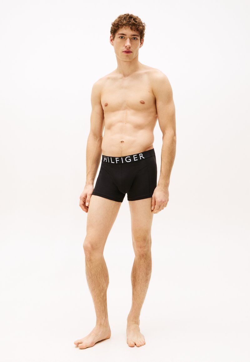 Fekete boxeralj egy fehér "HILFIGER" logóval a derékrészen, puha pamut keverékből készült. A modell mezítláb áll, egyenes testtartással.