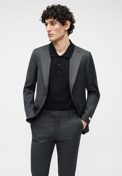 Άνδρας με σγουρά μαλλιά, φορώντας ένα σκούρο γκρι κοστούμι και μαύρο πόλο πουκάμισο, με τα χέρια στις τσέπες, κοιτάζοντας προς τα πλάγια, μπροστά σε απλό λευκό φόντο.