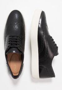 Chaussures en cuir noir avec détails brogue, dotées d'une semelle en caoutchouc blanche, d'un bout rond et d'un design traditionnel à lacets. Texture lisse et éclat subtil.