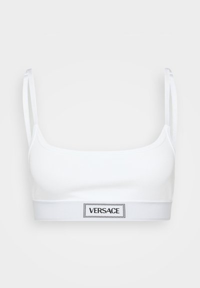 Balta kokvilnas bralette ar šaurām saitēm un plašu elastīgu joslu, kas rotāta ar melnu "VERSACE" logo taisnstūra formas uzlīmi.