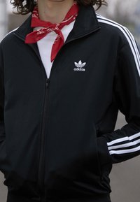 Svart zip-up jacka med tre vita ränder på ärmarna, med en Adidas-logotyp på vänster bröst. Bärs över en vit skjorta och en röd bandana.