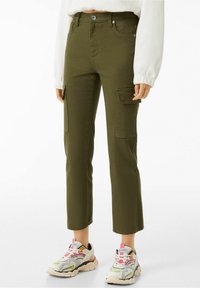 Osoba oblečená v olivových cargo kalhotách s postranními kapsami, bílém svetru a barevných robustních teniskách stojící na bílém pozadí.