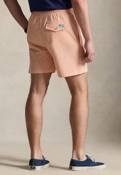 Ανδρικά ανοιχτό πορτοκαλί ριγέ σορτς με ελαστική μέση, πίσω τσέπη και λογότυπο. Φοριούνται με μπλε πάνινα παπούτσια.