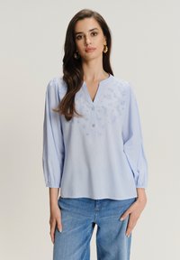 Blusa azzurro chiaro con scollatura a V, maniche a sbuffo, motivo floreale ricamato e chiusura con bottoni; indossata con jeans blu.