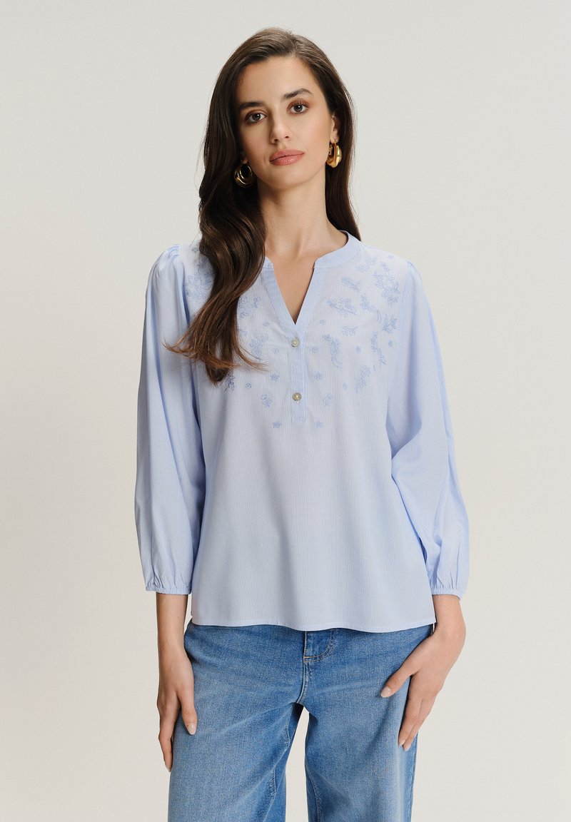 Blusa azzurro chiaro con scollatura a V, maniche a sbuffo, motivo floreale ricamato e chiusura con bottoni; indossata con jeans blu.