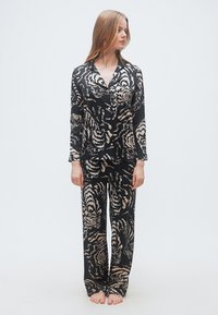 Γυναίκα που στέκεται ξυπόλυτη φορώντας μια σετ μαύρων πιτζάμων με μπεζ και λευκά μοτίβα τίγρης, κοιτώντας προς τα δεξιά της, σε λευκό φόντο.